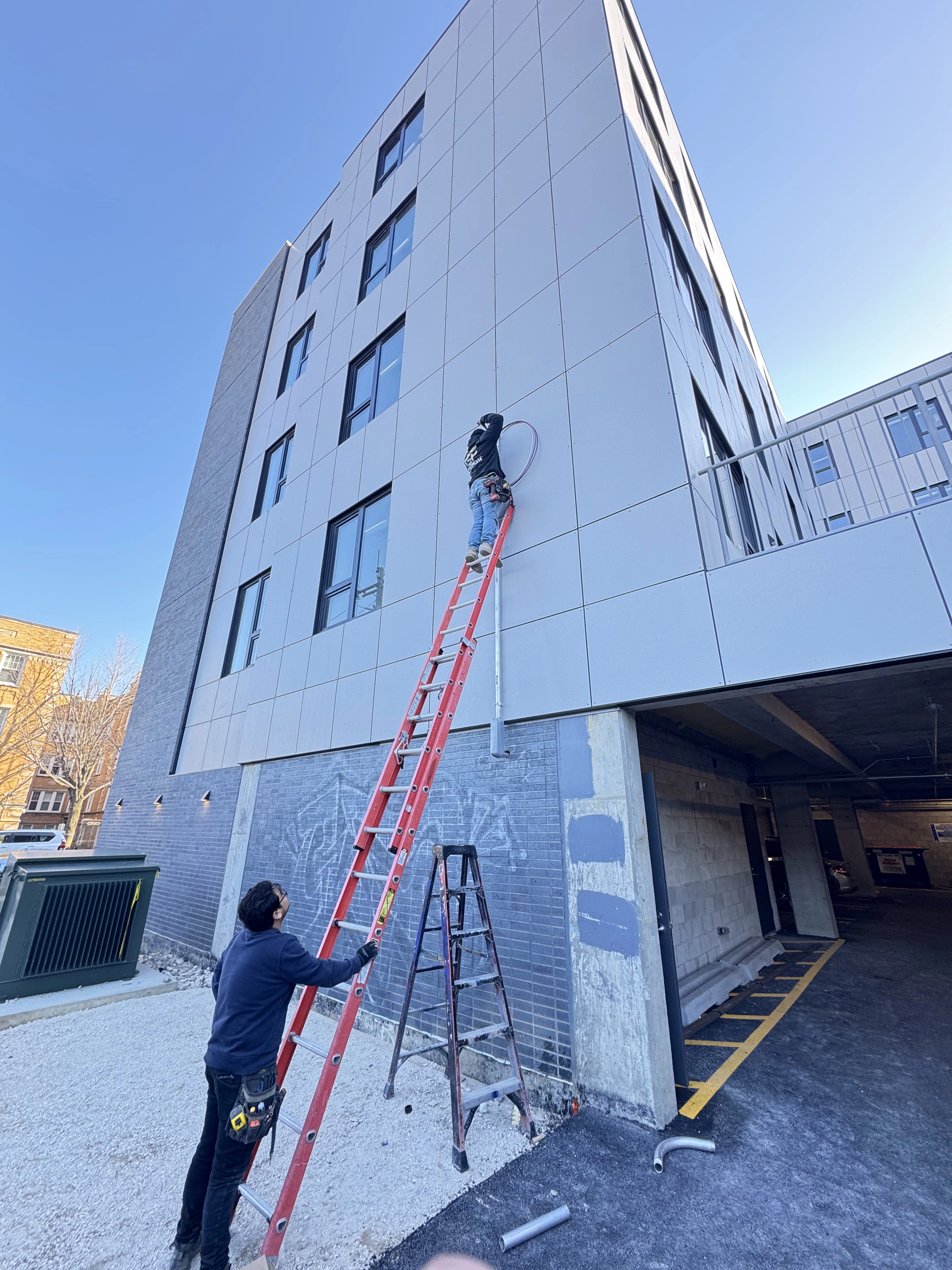 Two electricians alongside building, one on ladder working with wires
